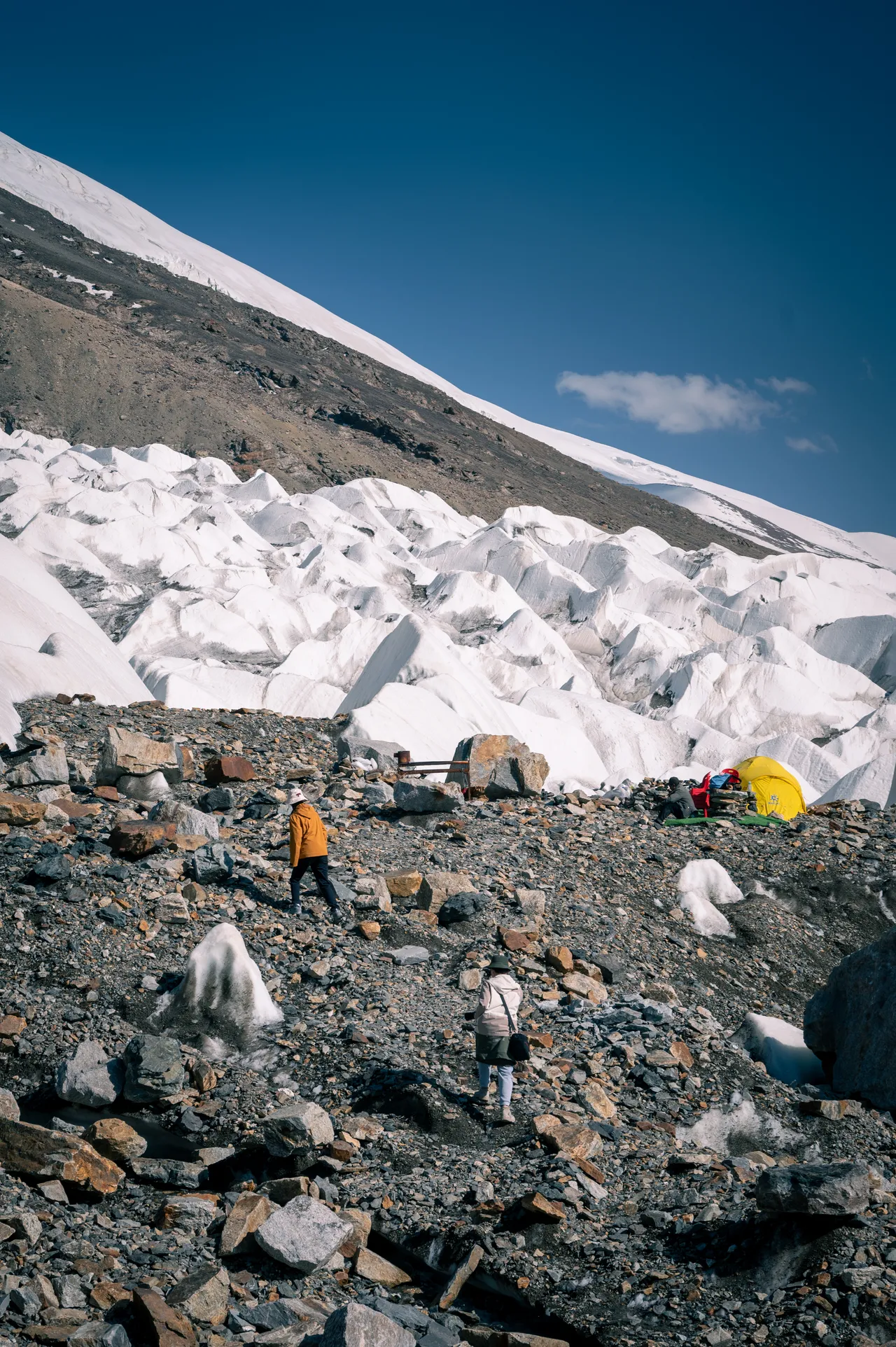 Hikers and a yellow tent beside glacier ice on a rocky mountainside
